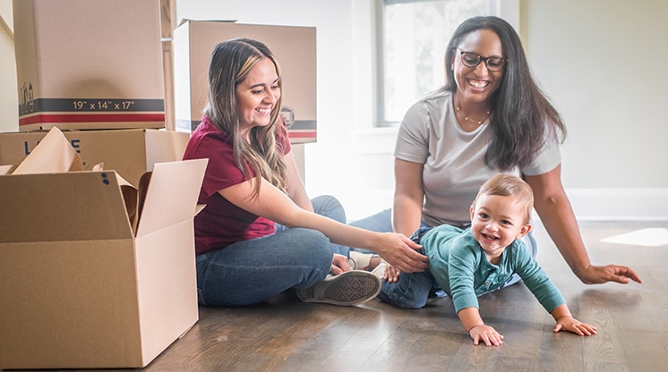 two women watch baby crawl on floor with moving boxes